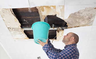 a man is using a bucket to capture water above a pipe break in the ceiling.