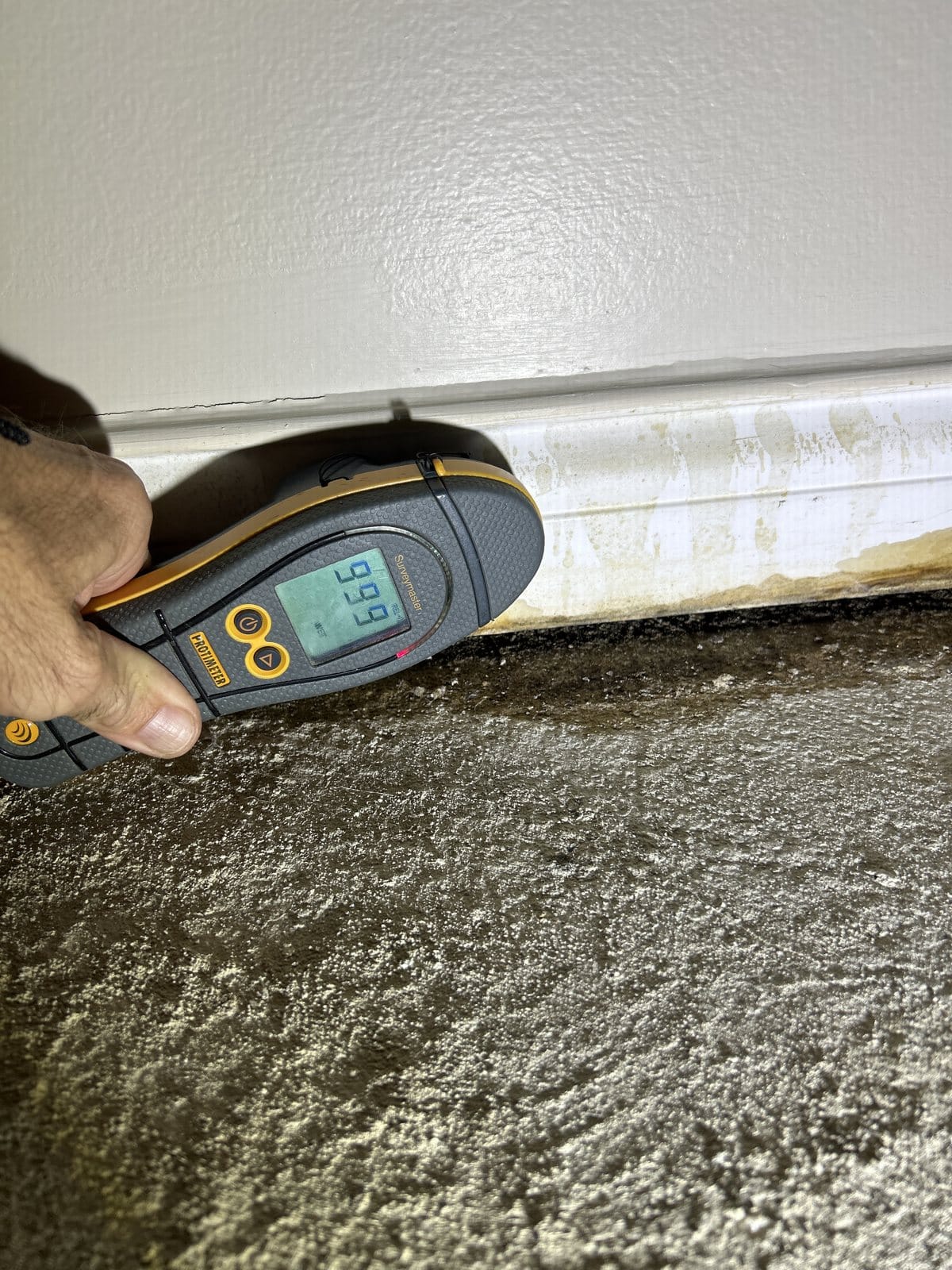 Moisture meter reading 99.9 on concrete wall showing efflorescence and discoloration—diagnostic phase of water damage assessment
